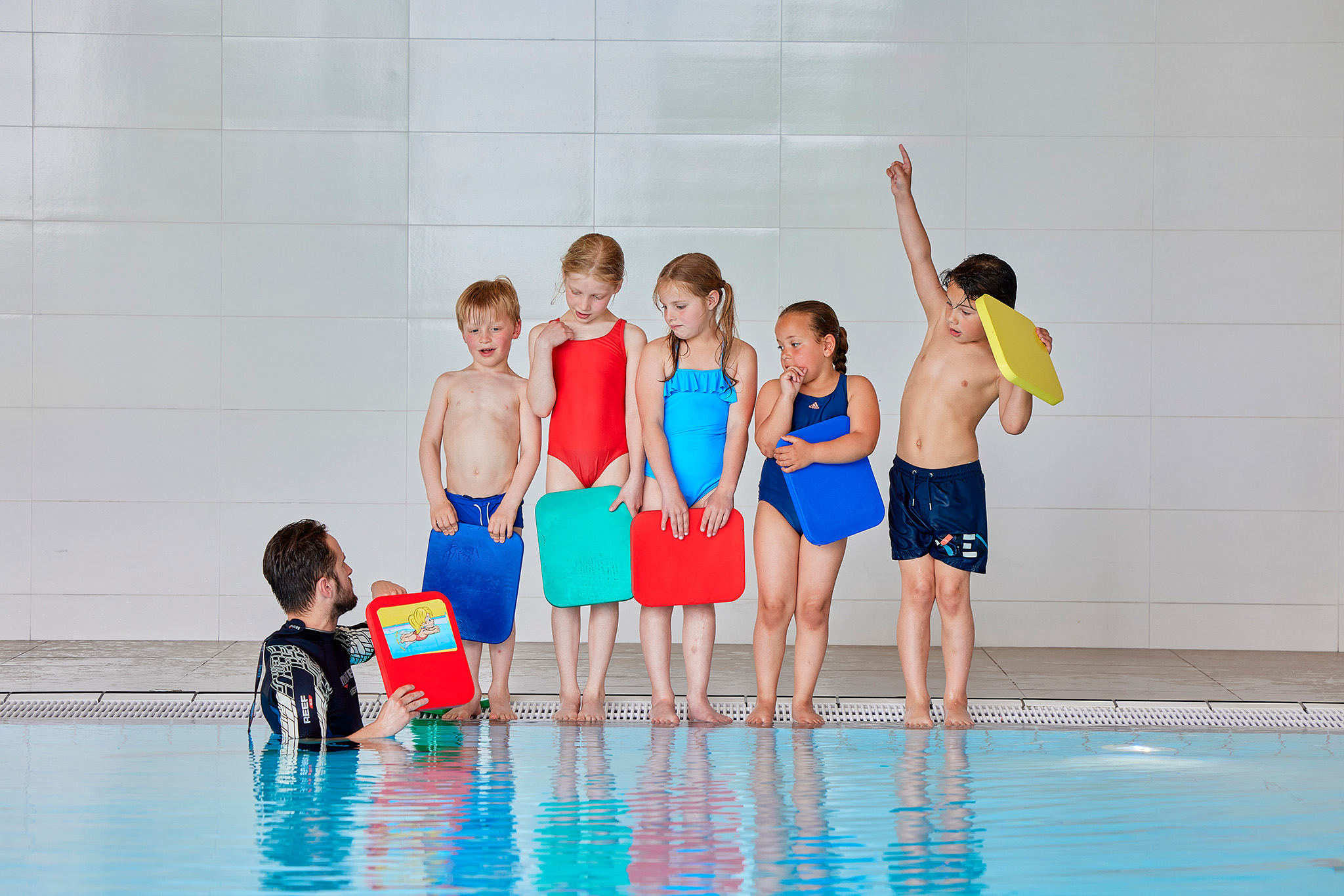 Vijf kinderen staan op de rand van het zwembad tijdens de zwemles bij Optisport, ze luisteren naar de instructeur die ze vanuit het water instructies geeft. 