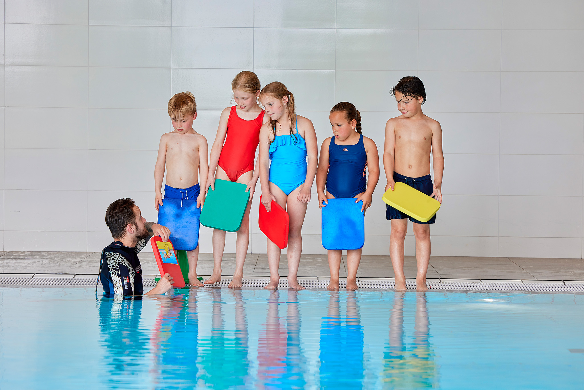 Vijf kinderen staan op de rand van het zwembad van Optisport en luisteren geconcentreerd naar de instructeur, die vanuit de Zed en Sop zwemlesmethode instructies geeft. 